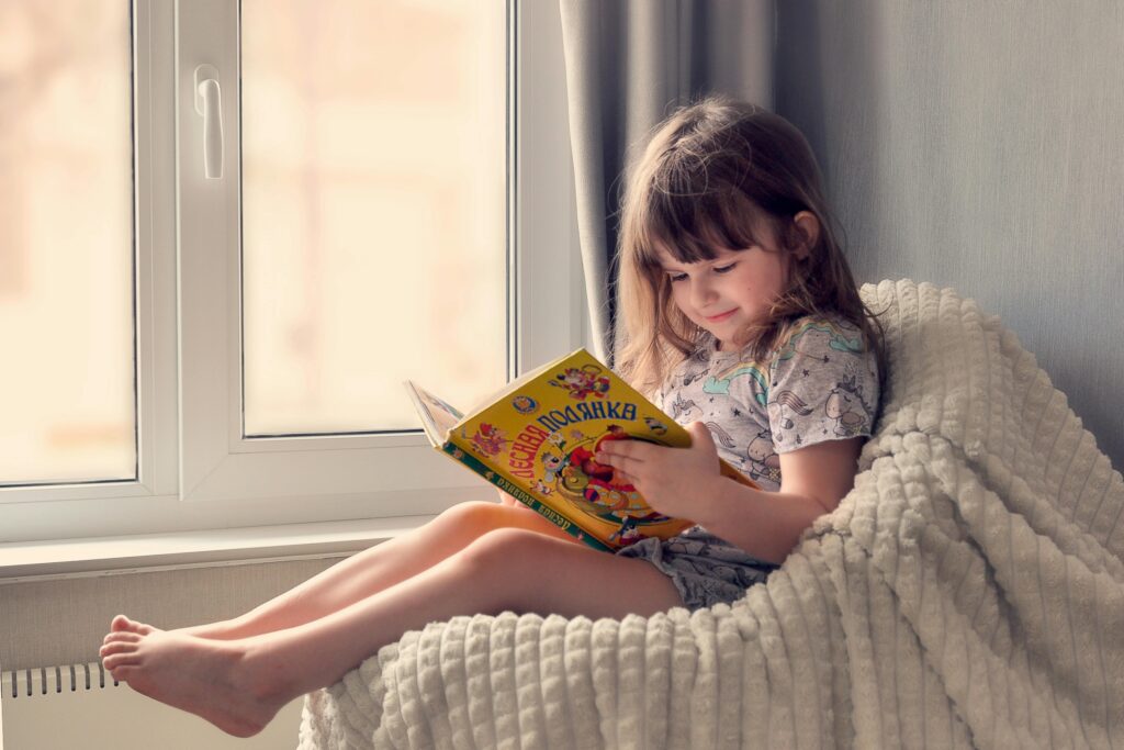 A young girl reads a colorful book by the window, embracing cozy and peaceful leisure time indoors.