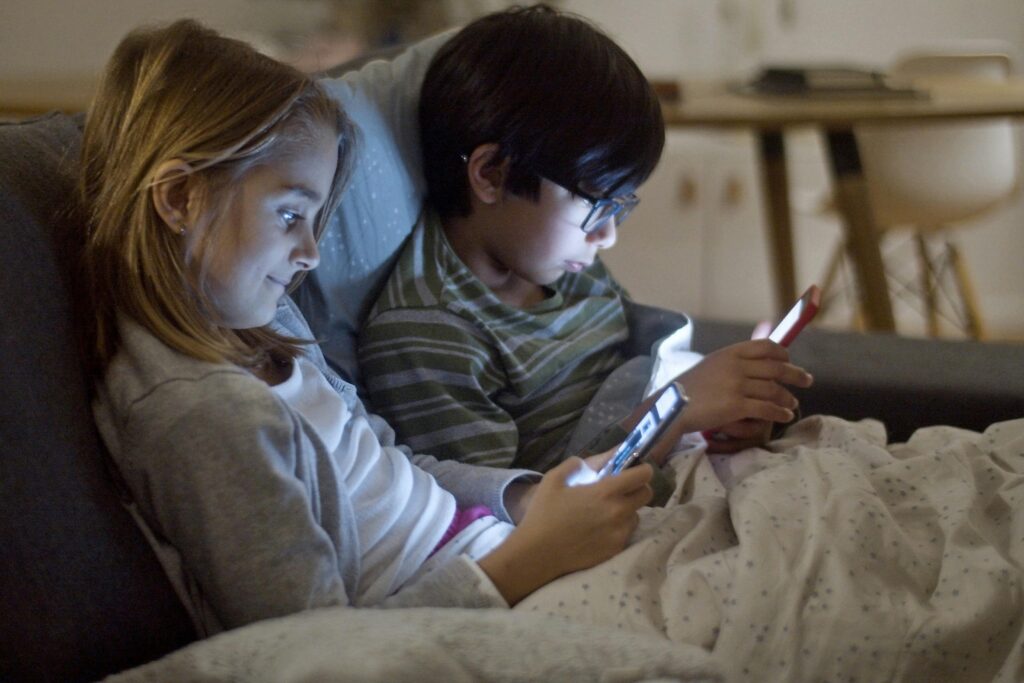 Two children using smartphones at night, indoors, under blankets.